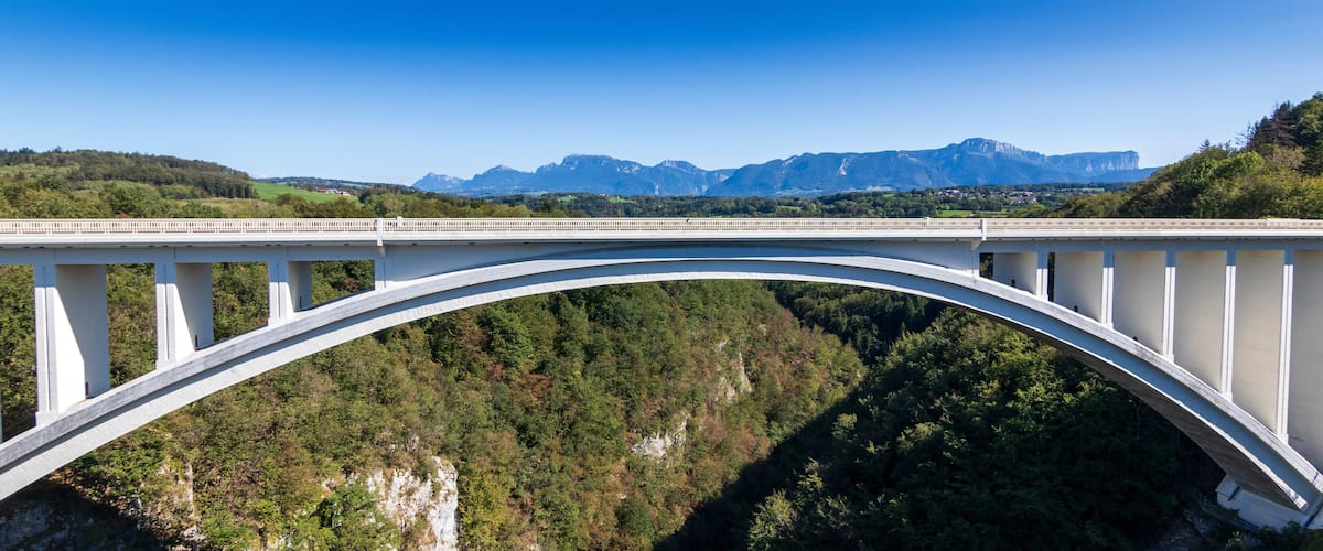 A white bridge spanning a deep gorge with forested hills and mountains in the background.