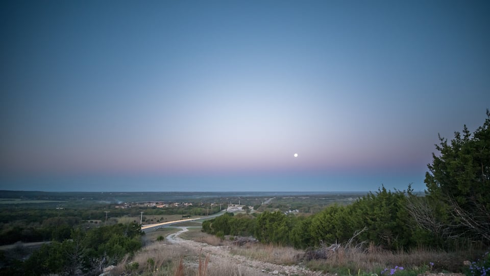 A landscape with a road, trees, and a moon in the sky.