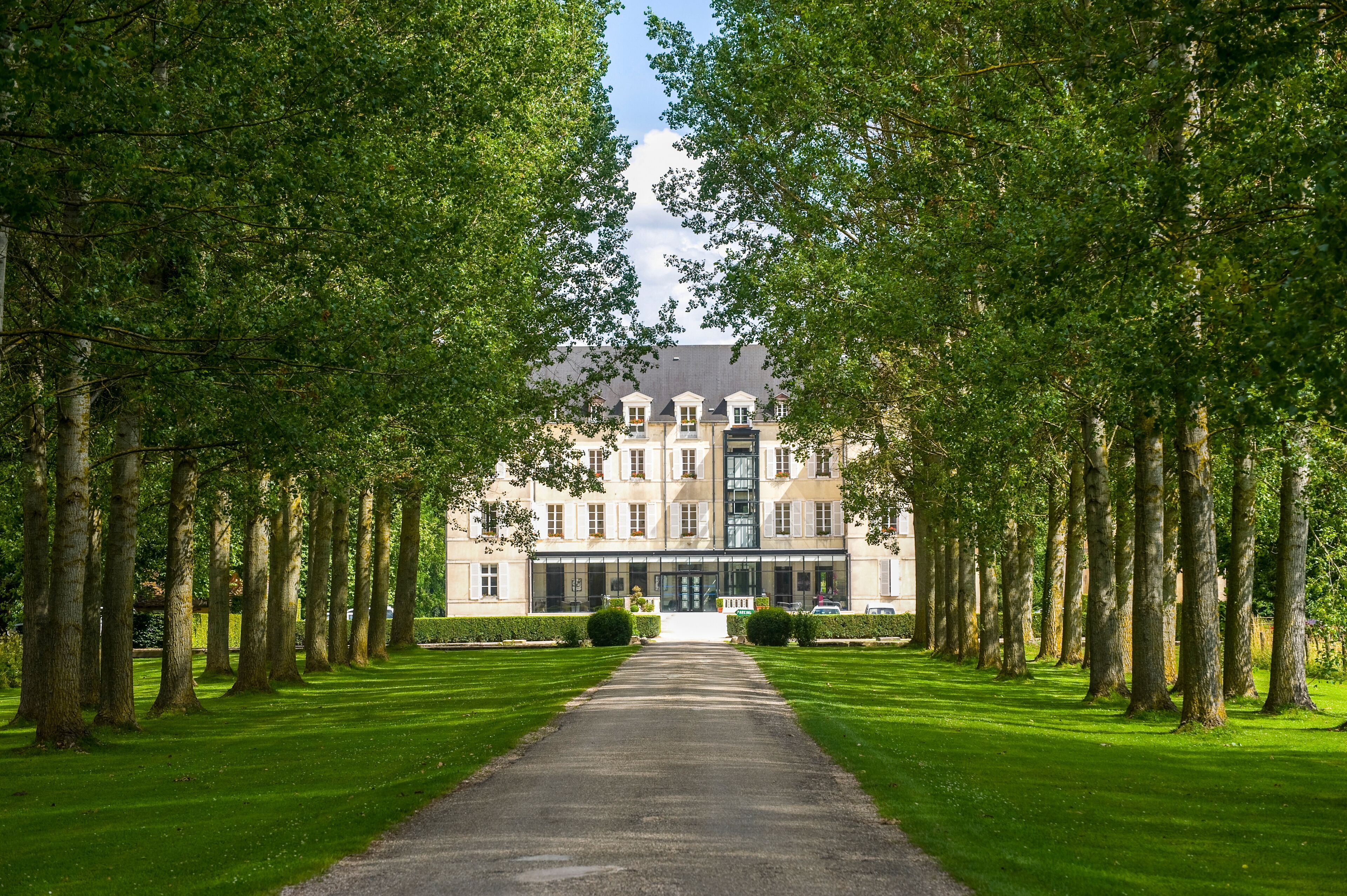 A grand building with a symmetrical facade, flanked by rows of tall trees and a well-maintained lawn.
