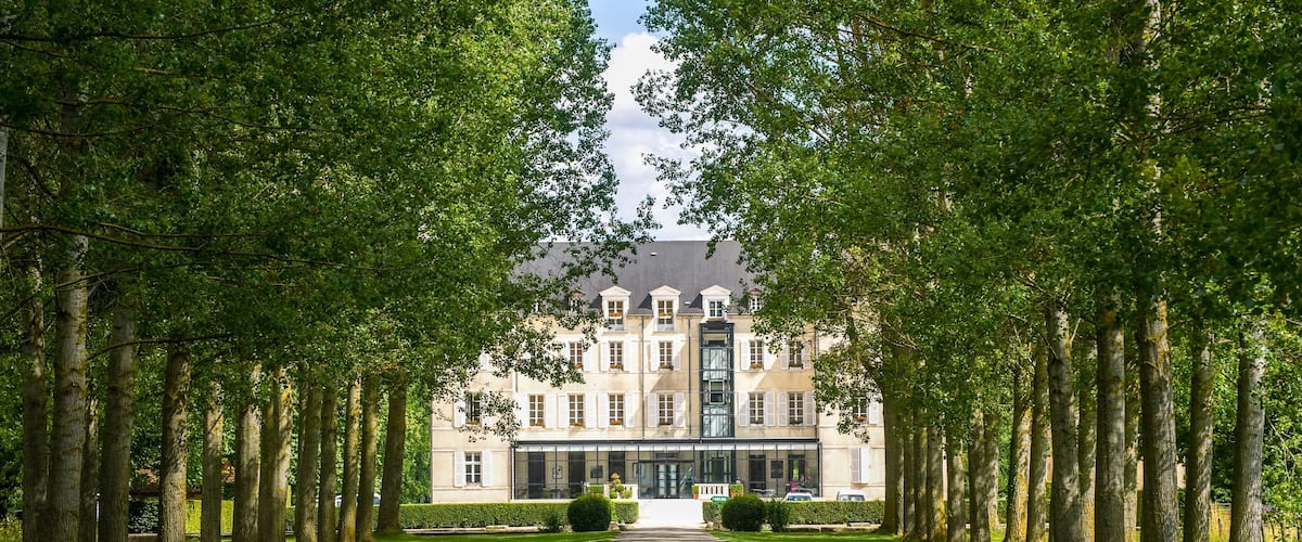 A grand building with a symmetrical facade, flanked by rows of tall trees and a well-maintained lawn.