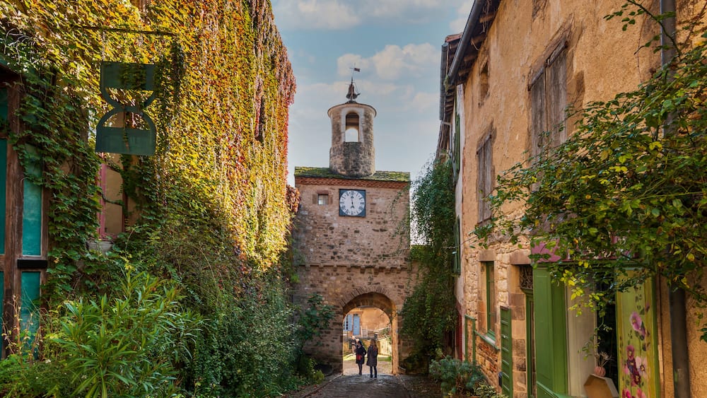Une ruelle étroite pavée, bordée d’immeubles recouverts de lierre, qui mène à une tour surmontée d’une horloge.