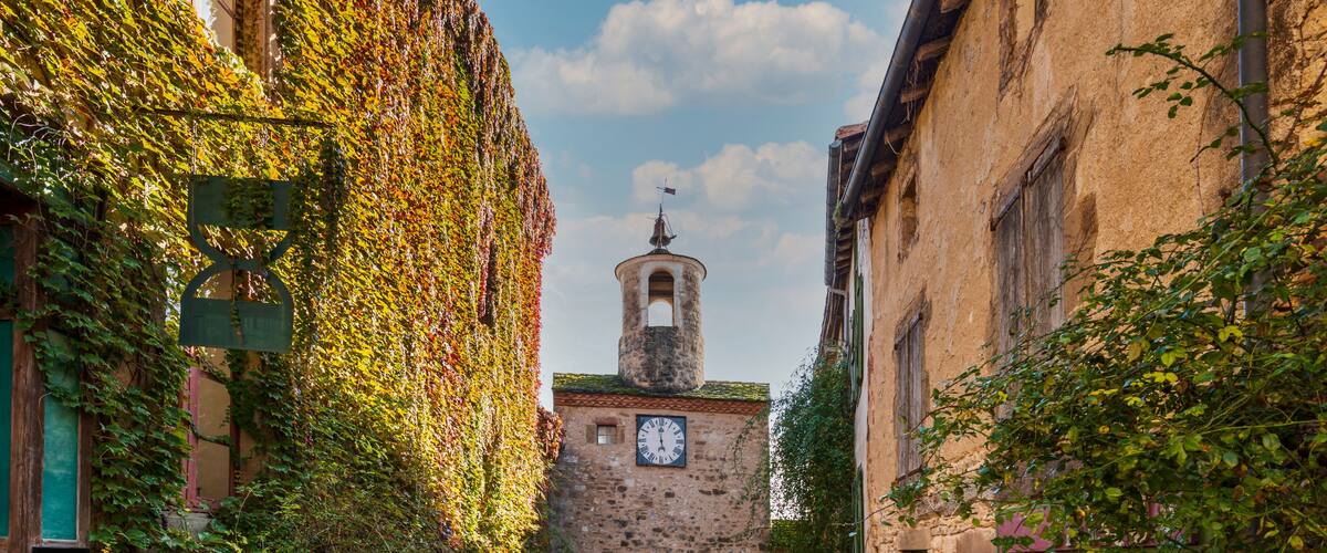 Un estrecho callejón adoquinado flanqueado por edificios cubiertos de hiedra, que conduce a una torre con un reloj.