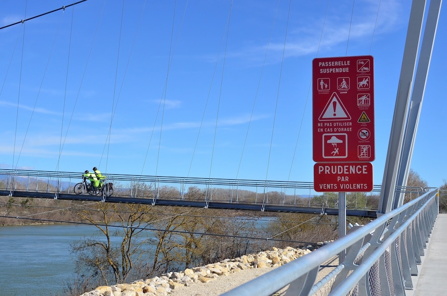 Un puente colgante con un cartel que advierte sobre un pasillo peatonal suspendido y recomienda precaución debido a fuertes vientos.