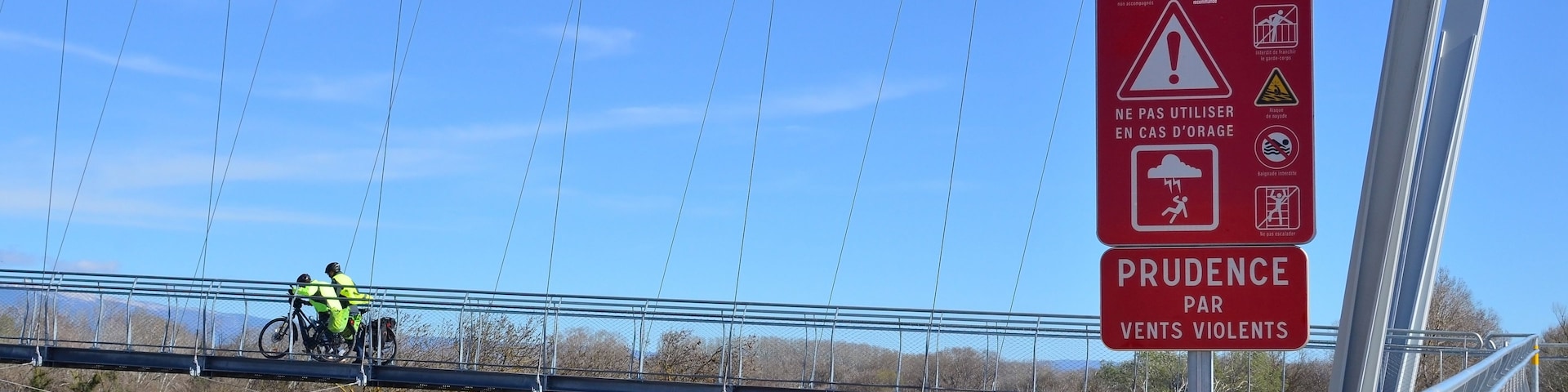A suspension bridge with a sign warning of a suspended pedestrian walkway and advising caution due to strong winds.