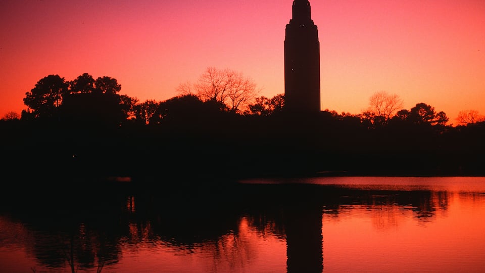 A silhouette of a tall tower against a sunset sky, with trees reflected in a body of water.