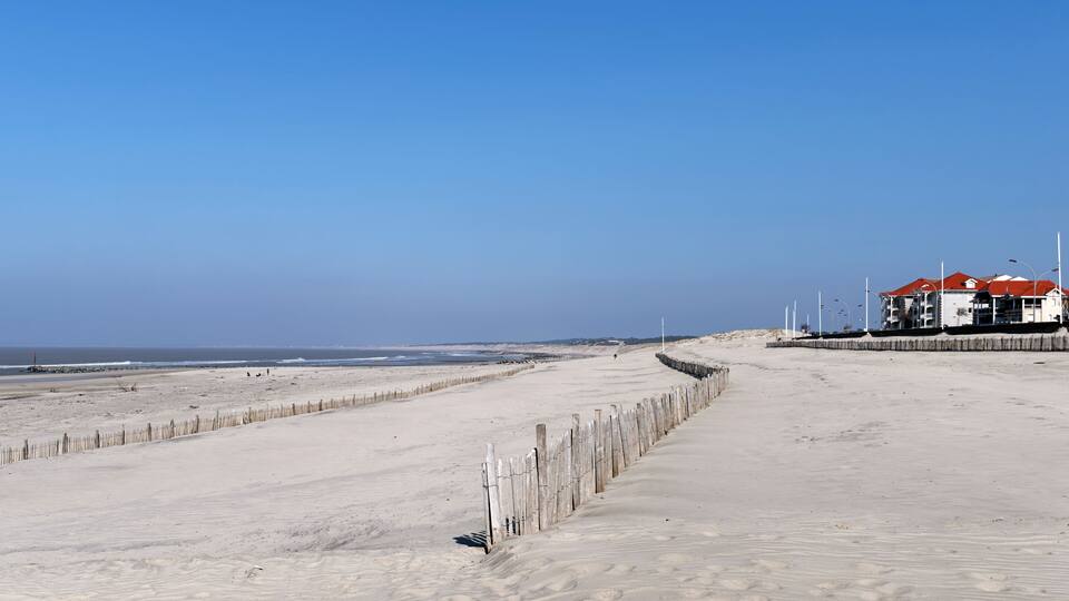 A sandy beach with a wooden fence, a building with a red roof, and the ocean in the background.