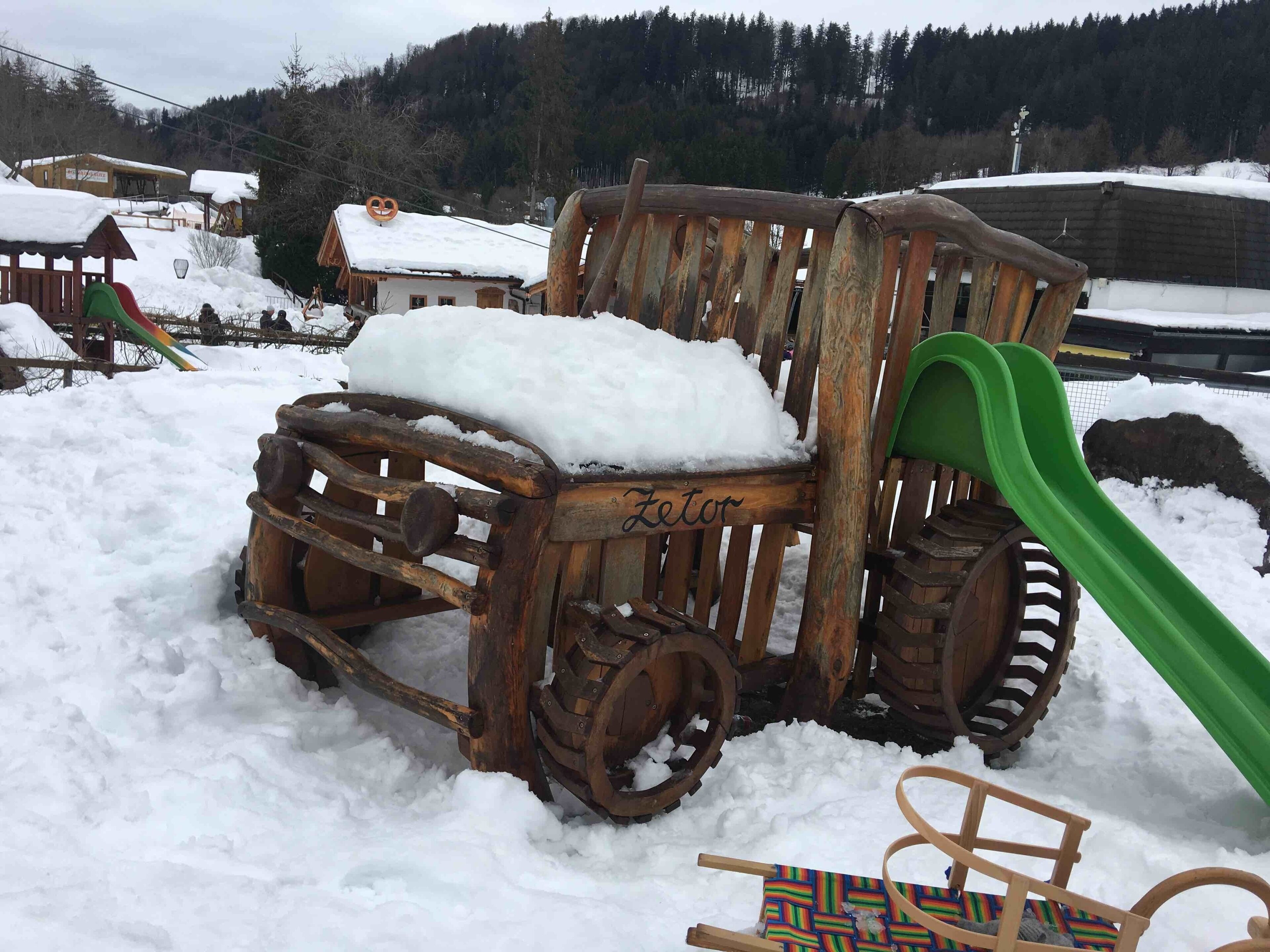 A wooden sled with a green slide, a wooden chair, and a colorful woven seat in a snowy outdoor setting.
