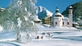 A snow-covered landscape with a church, a mountain, and a few skiers.