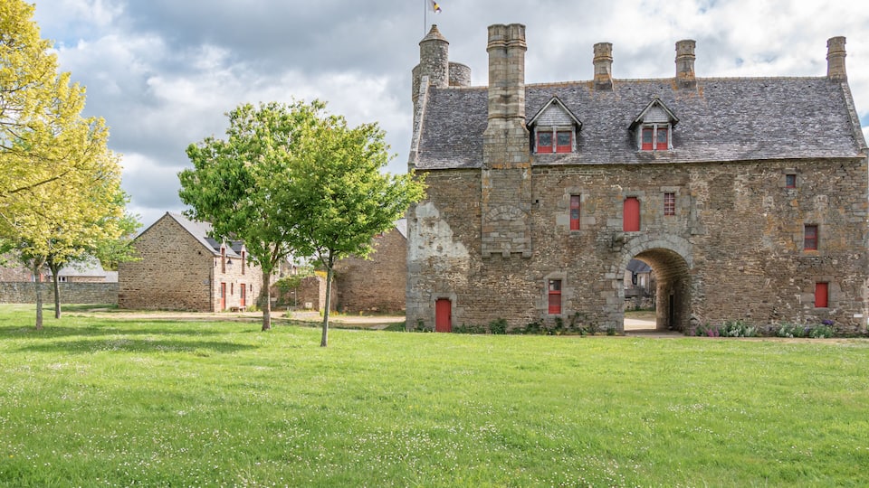 A historic stone building with red shutters, a large tree in front, and a grassy field.