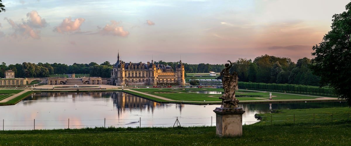 A grand palace with a large lake, manicured gardens, and a statue in the foreground.