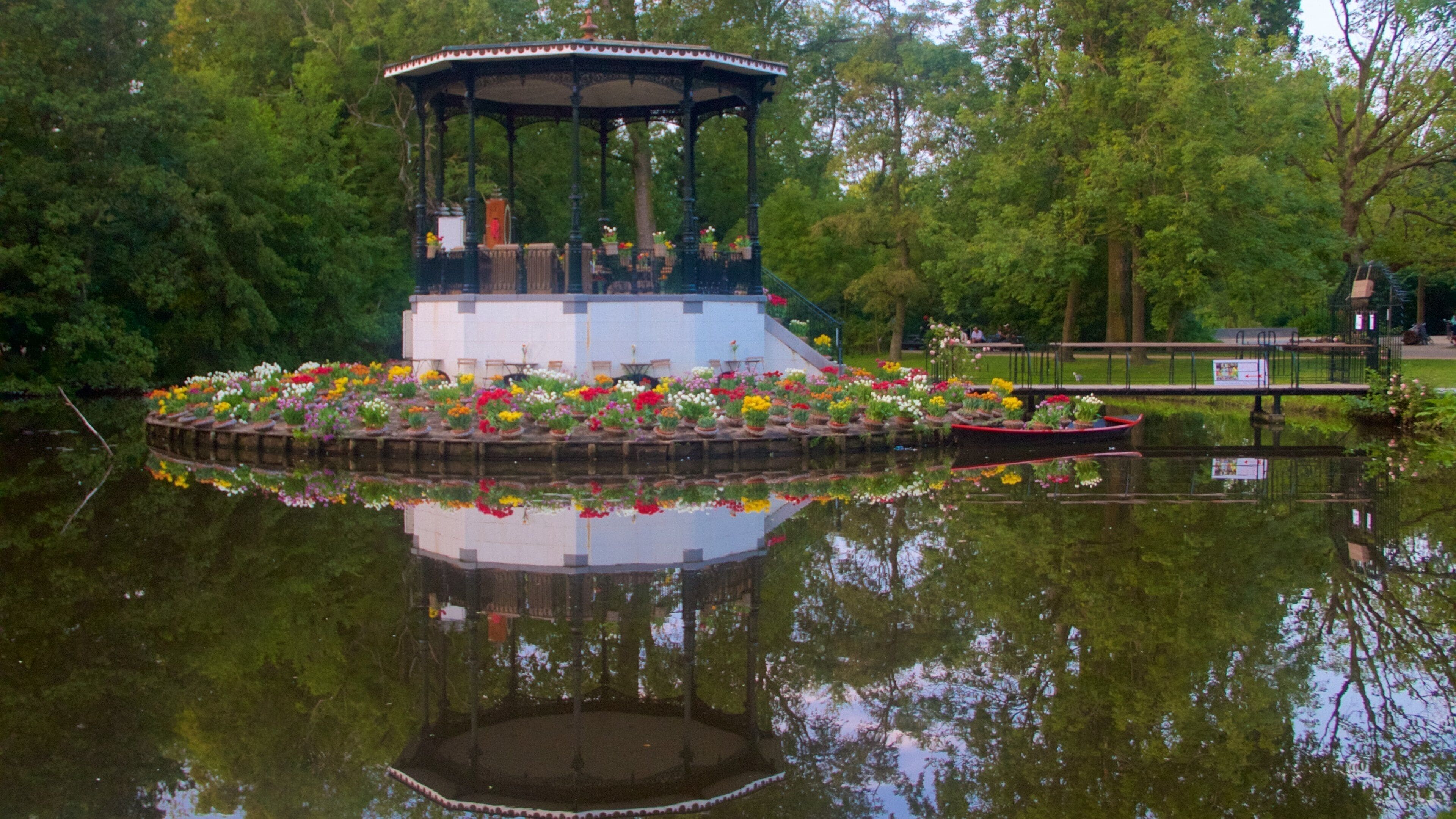 A gazebo with a flower garden in the middle of a pond, surrounded by trees.