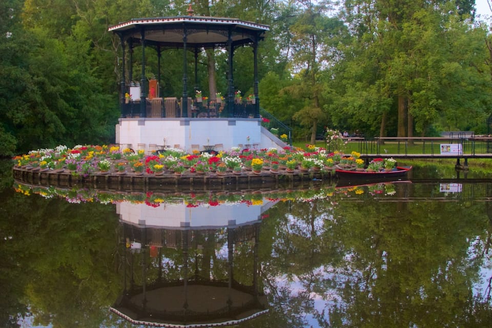 A gazebo with a flower garden in the middle of a pond, surrounded by trees.