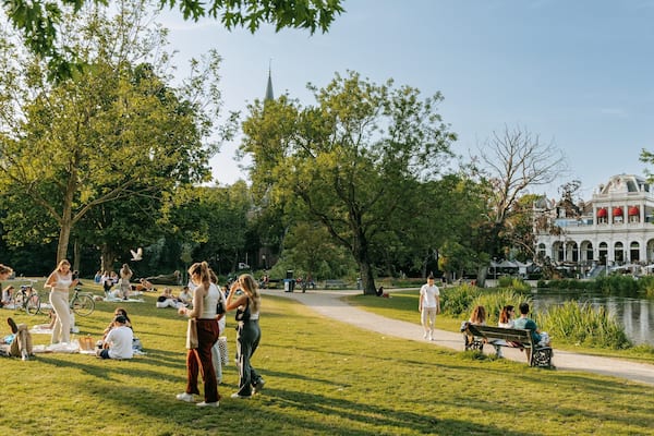 Ein Park mit Menschen, die auf der Wiese entspannen, einem Teich und einem historischen Gebäude im Hintergrund.