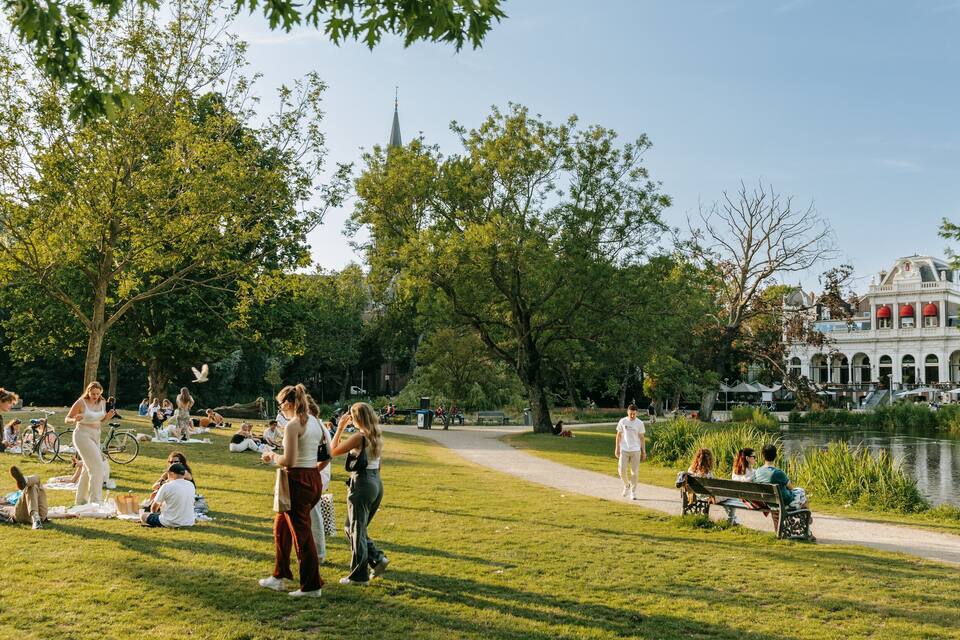 Un parque con gente descansando sobre el césped, un estanque y un edificio histórico al fondo.