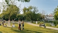 Un parc amb gent relaxant-se sobre l'herba, un estany i un edifici històric al fons.