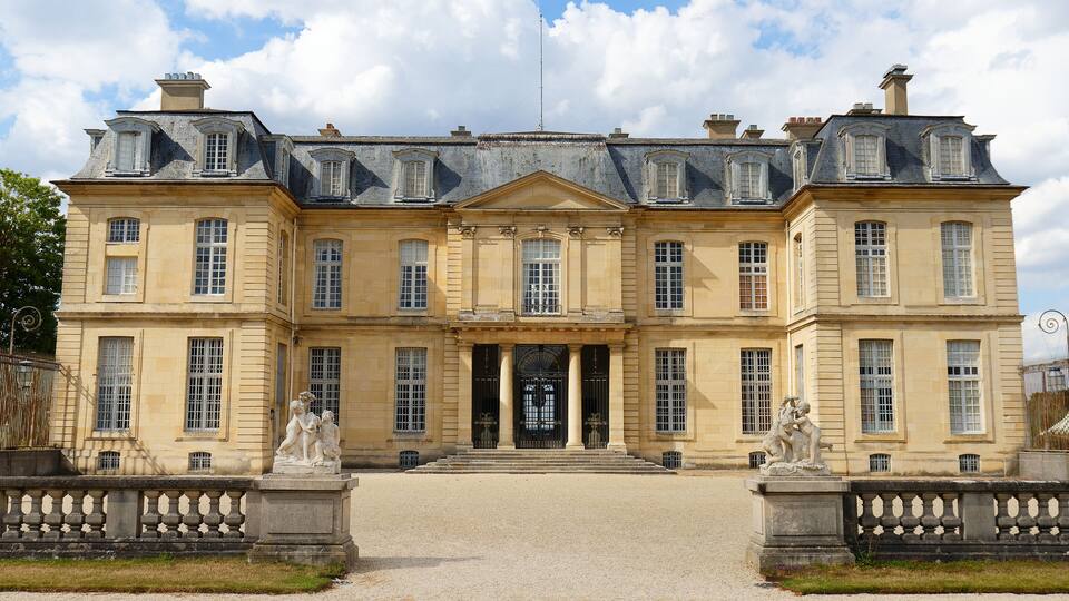 A large, historic building with a symmetrical facade, large windows, and statues at the entrance.