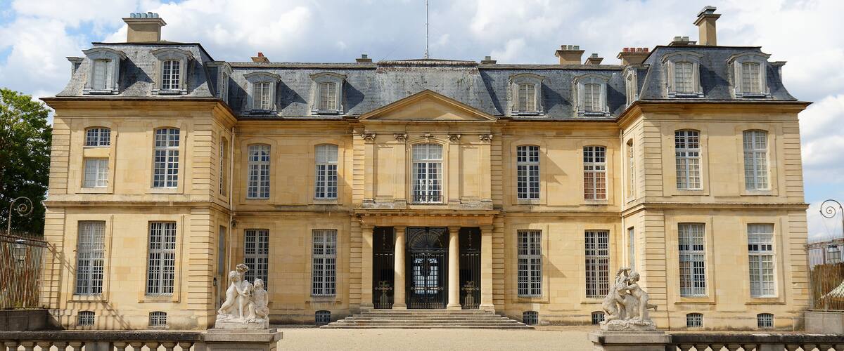 A large, historic building with a symmetrical facade, large windows, and statues at the entrance.