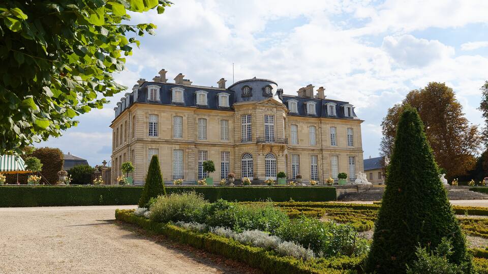 A large classical building with a symmetrical facade, surrounded by manicured gardens and a gravel path.