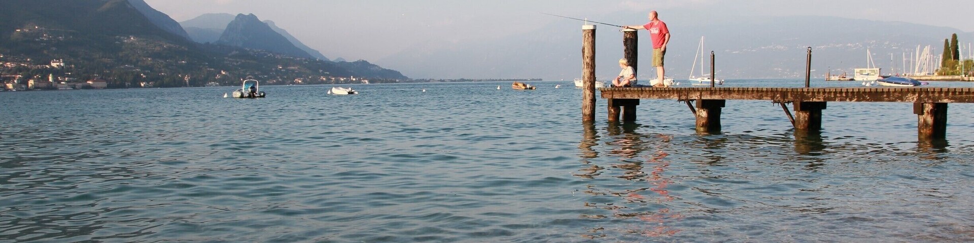 A pier extending into a calm lake with boats, mountains, and a clear sky.