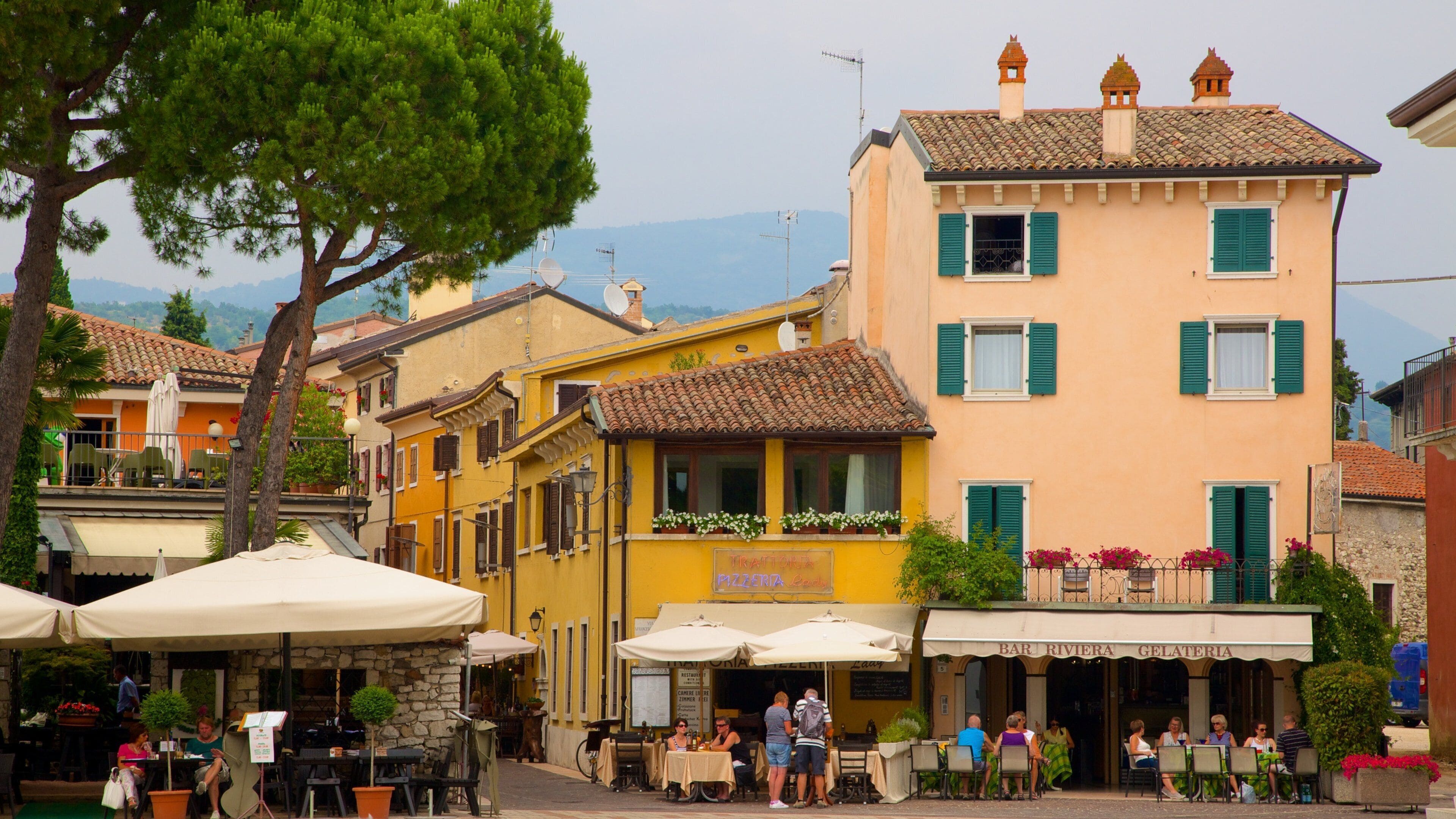 A quaint European street with colorful buildings, outdoor seating, and a mountainous backdrop.