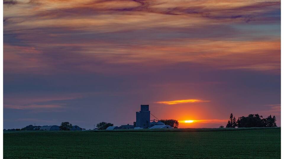 A sunset over a rural landscape with a silo and farm buildings.