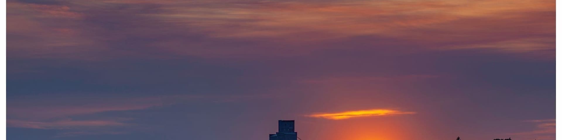 A sunset over a rural landscape with a silo and farm buildings.