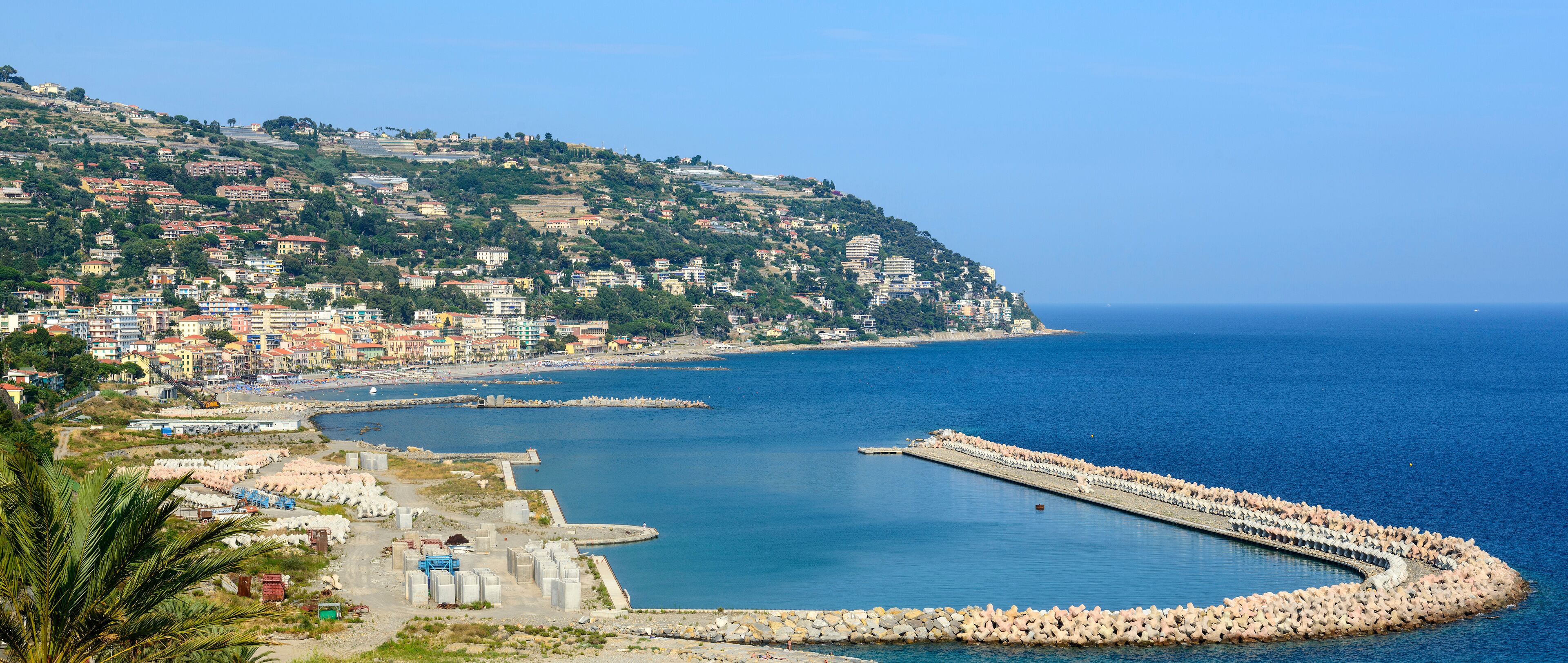 A coastal town with a harbor, a rocky breakwater, and a hillside with buildings.