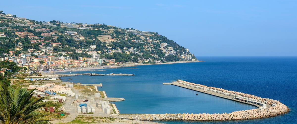 A coastal town with a harbor, a rocky breakwater, and a hillside with buildings.