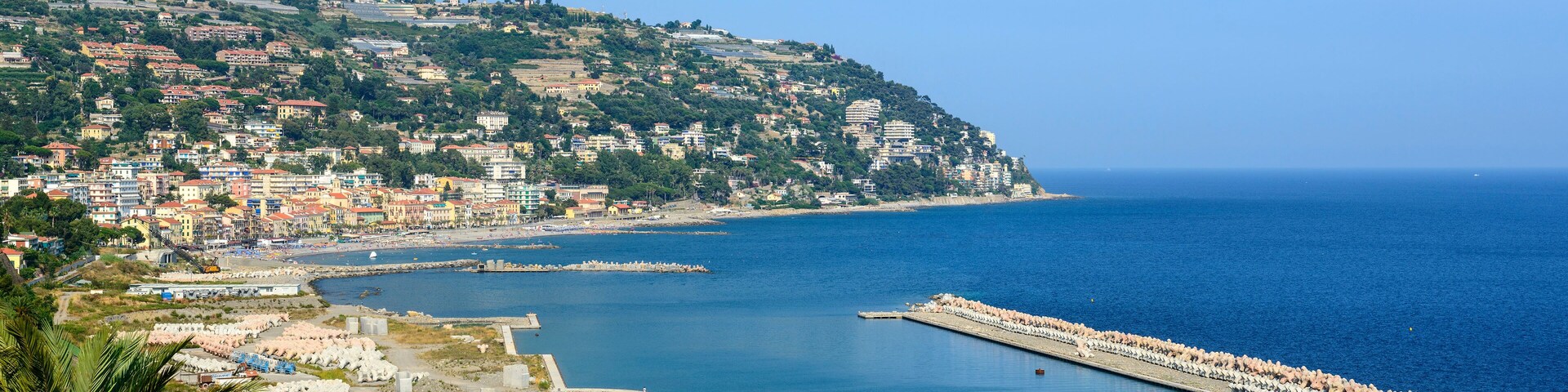 A coastal town with a harbor, a rocky breakwater, and a hillside with buildings.