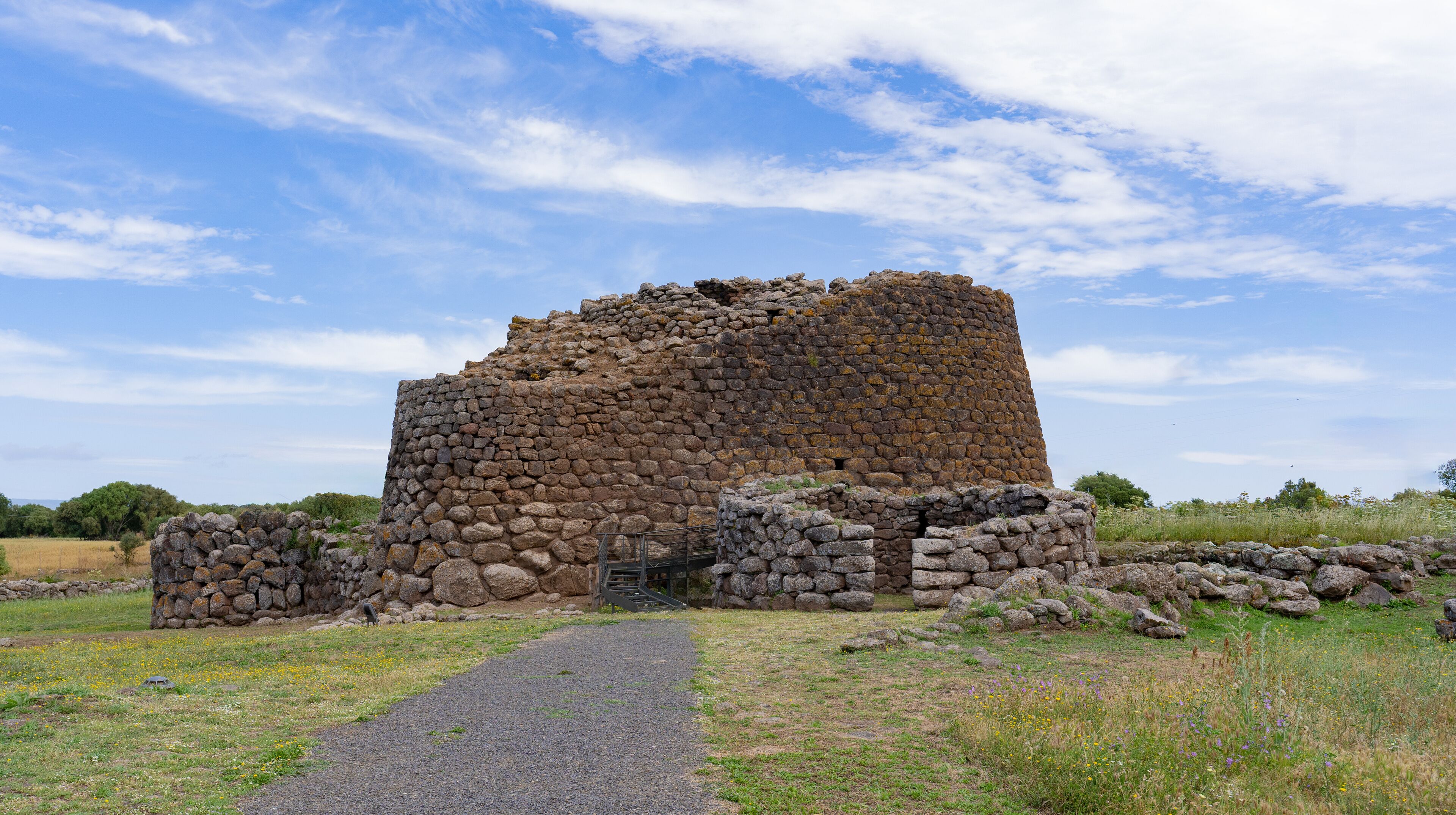 A stone ruin with a gravel path leading to it, surrounded by grass and wildflowers.