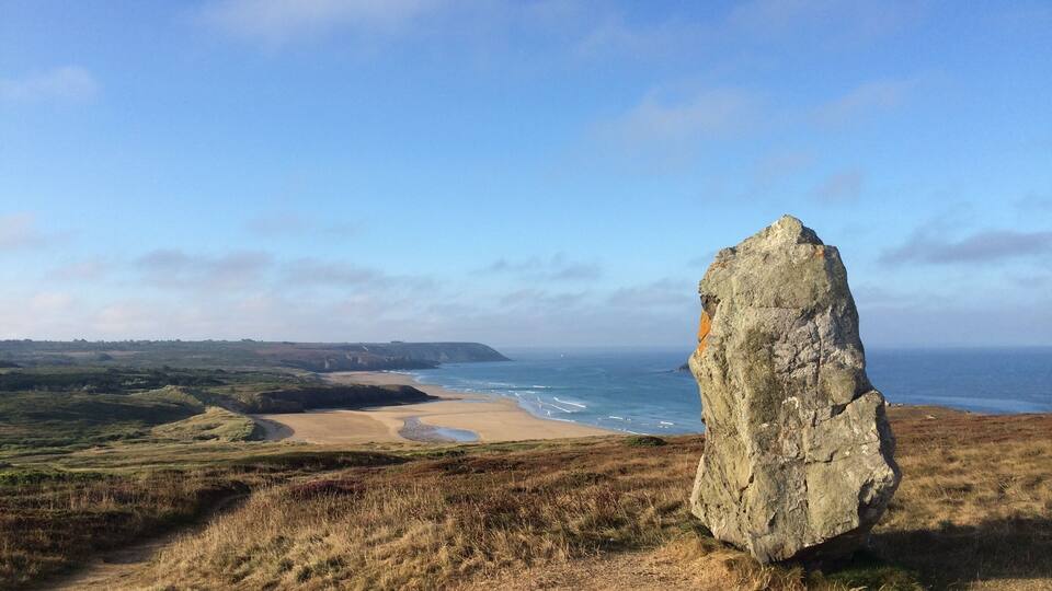 A coastal landscape with a prominent rock formation, sandy beach, and rolling hills.