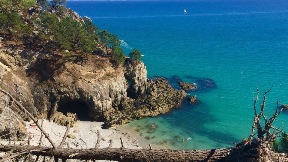 A coastal landscape with rocky cliffs, clear turquoise waters, and a sailboat in the distance.