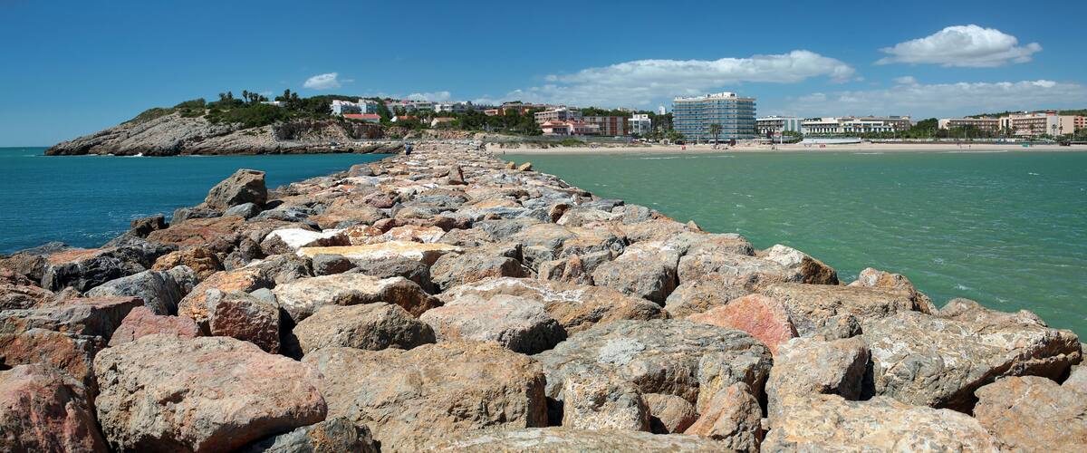 Una costa rocosa con mar azul cristalino, una playa arenosa y edificios al fondo.