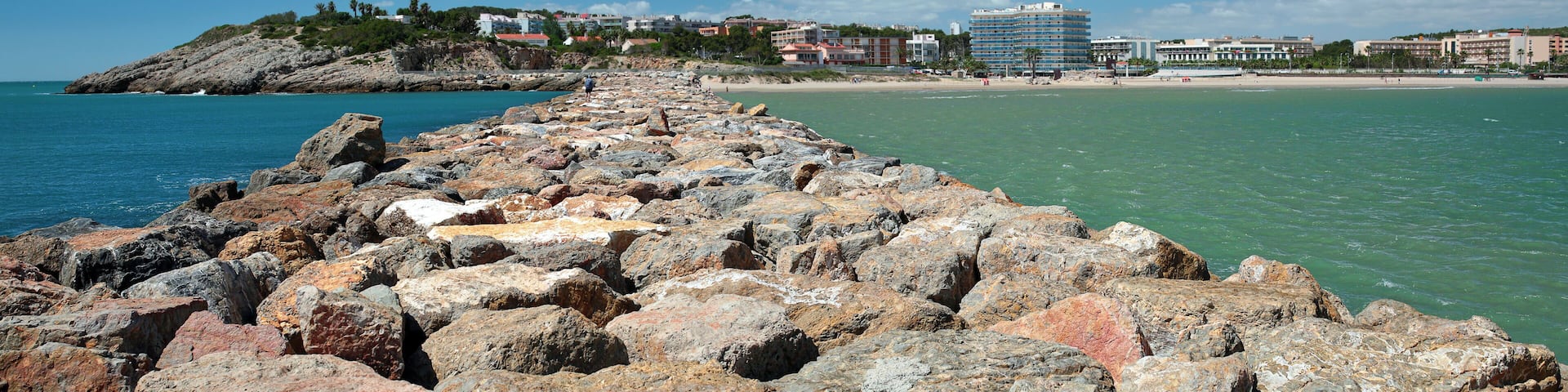 A rocky shoreline with a clear blue sea, a sandy beach, and buildings in the background.