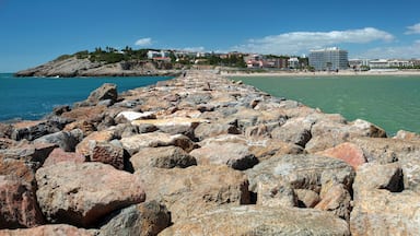 A rocky shoreline with a clear blue sea, a sandy beach, and buildings in the background.