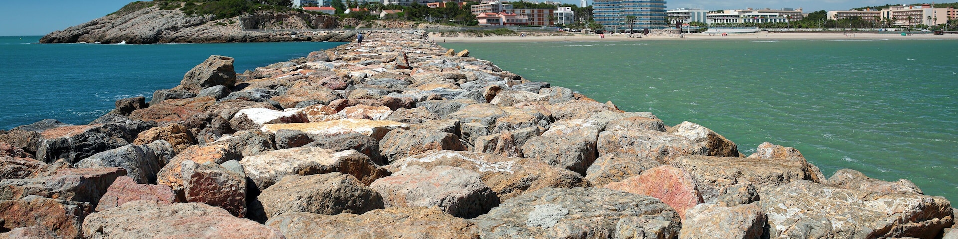 Una costa rocosa con mar azul cristalino, una playa arenosa y edificios al fondo.