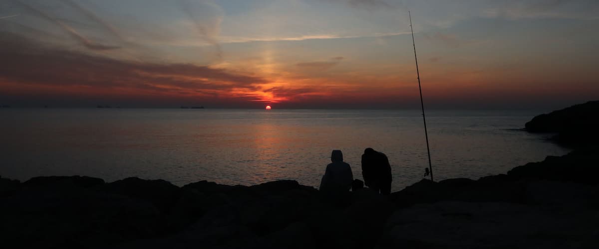 A sunset over a calm sea with silhouettes of two people and a fishing rod on the shore.