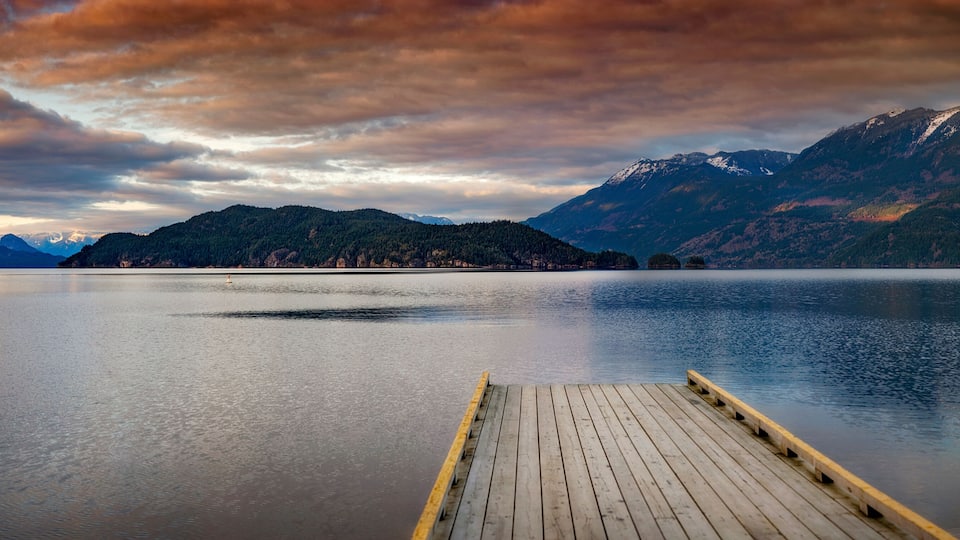 A calm lake with a wooden dock extending into it, surrounded by mountains and a cloudy sky.