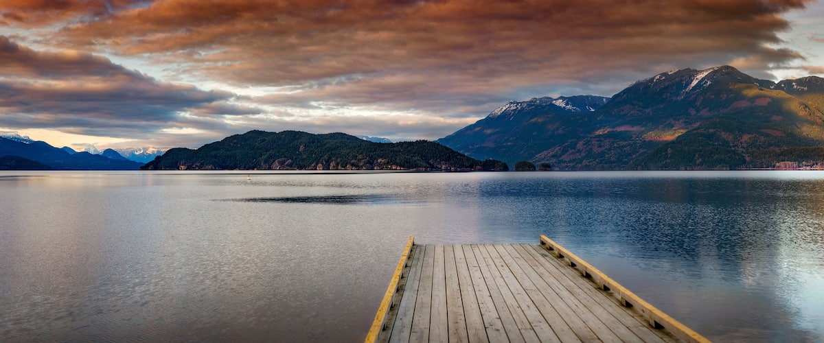 A calm lake with a wooden dock extending into it, surrounded by mountains and a cloudy sky.