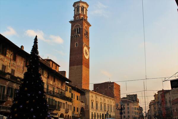Une haute tour de l’horloge se dresse sur une place de la ville, entourée de bâtiments historiques et d’un sapin de Noël décoré.