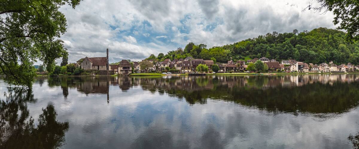 A calm lake reflecting houses and trees, surrounded by lush greenery and a cloudy sky.