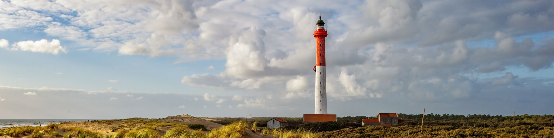A lighthouse on a sandy dune with grass, a cloudy sky, and a distant view of the ocean.