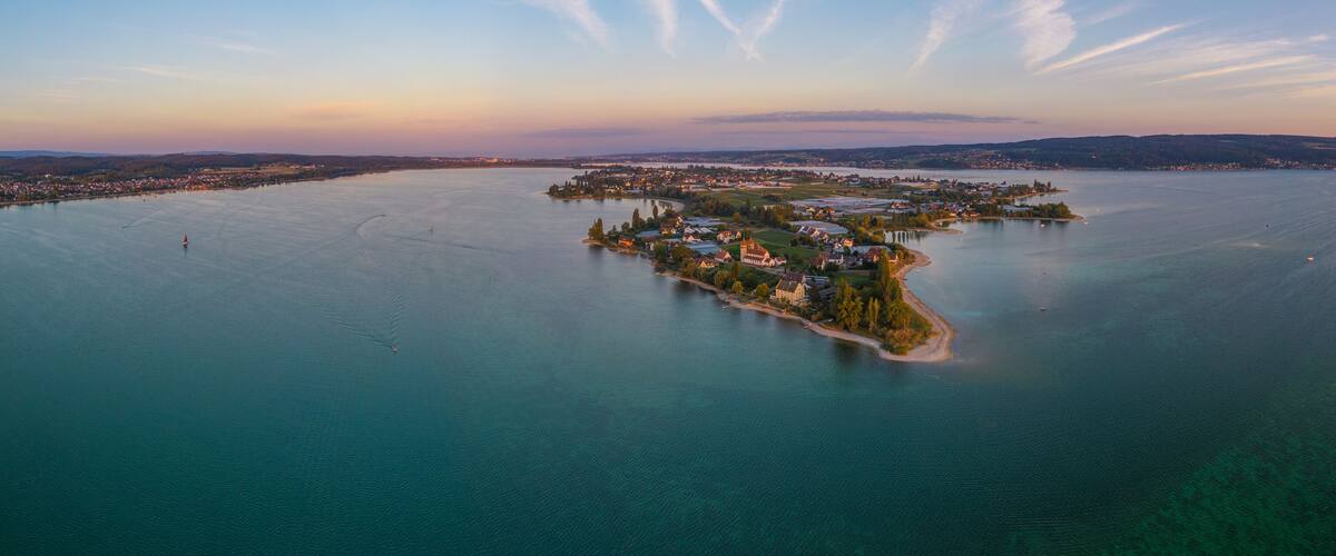 An aerial view of a small island with buildings and a lighthouse, surrounded by calm water and distant hills.