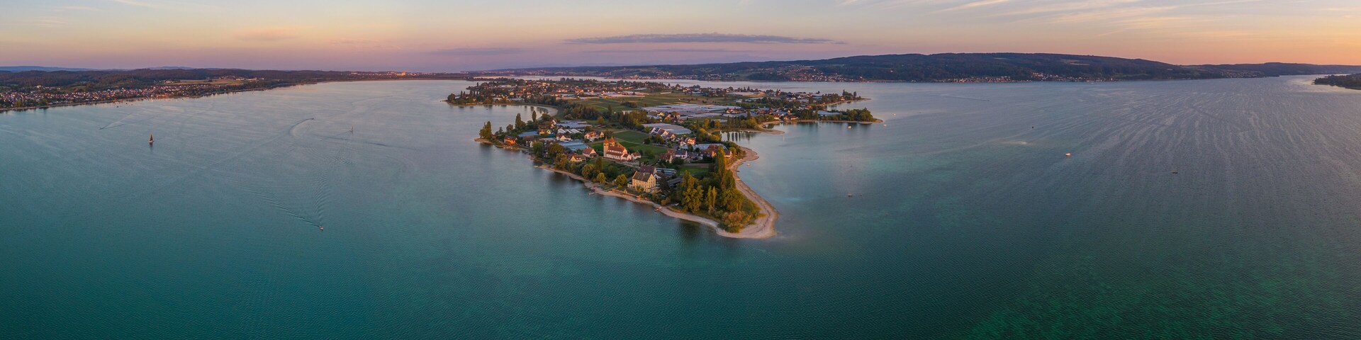 An aerial view of a small island with buildings and a lighthouse, surrounded by calm water and distant hills.