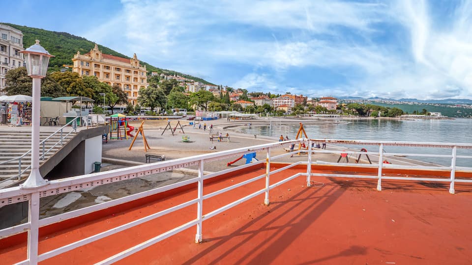 A waterfront promenade with playgrounds, a white railing, and buildings in the background.