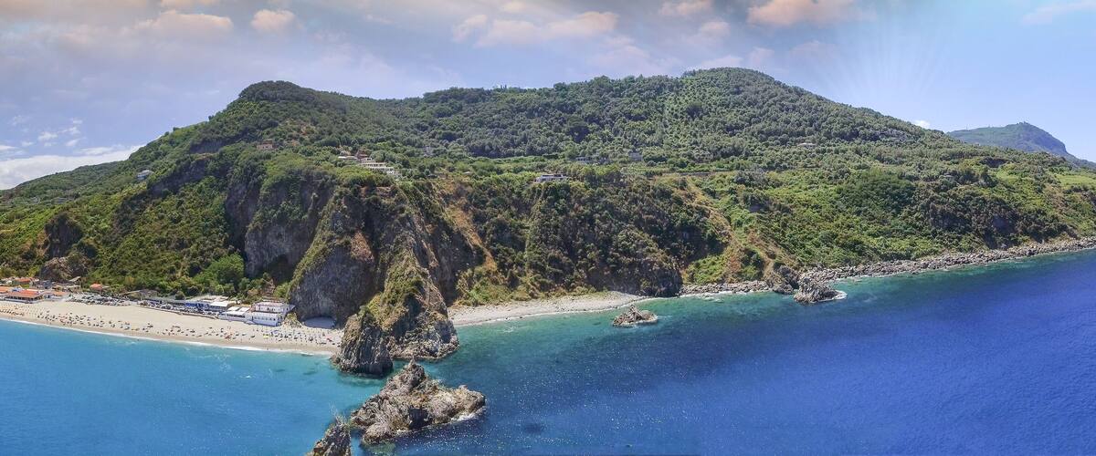 A coastal landscape with a mountainous island, clear blue sea, and a beach with buildings.