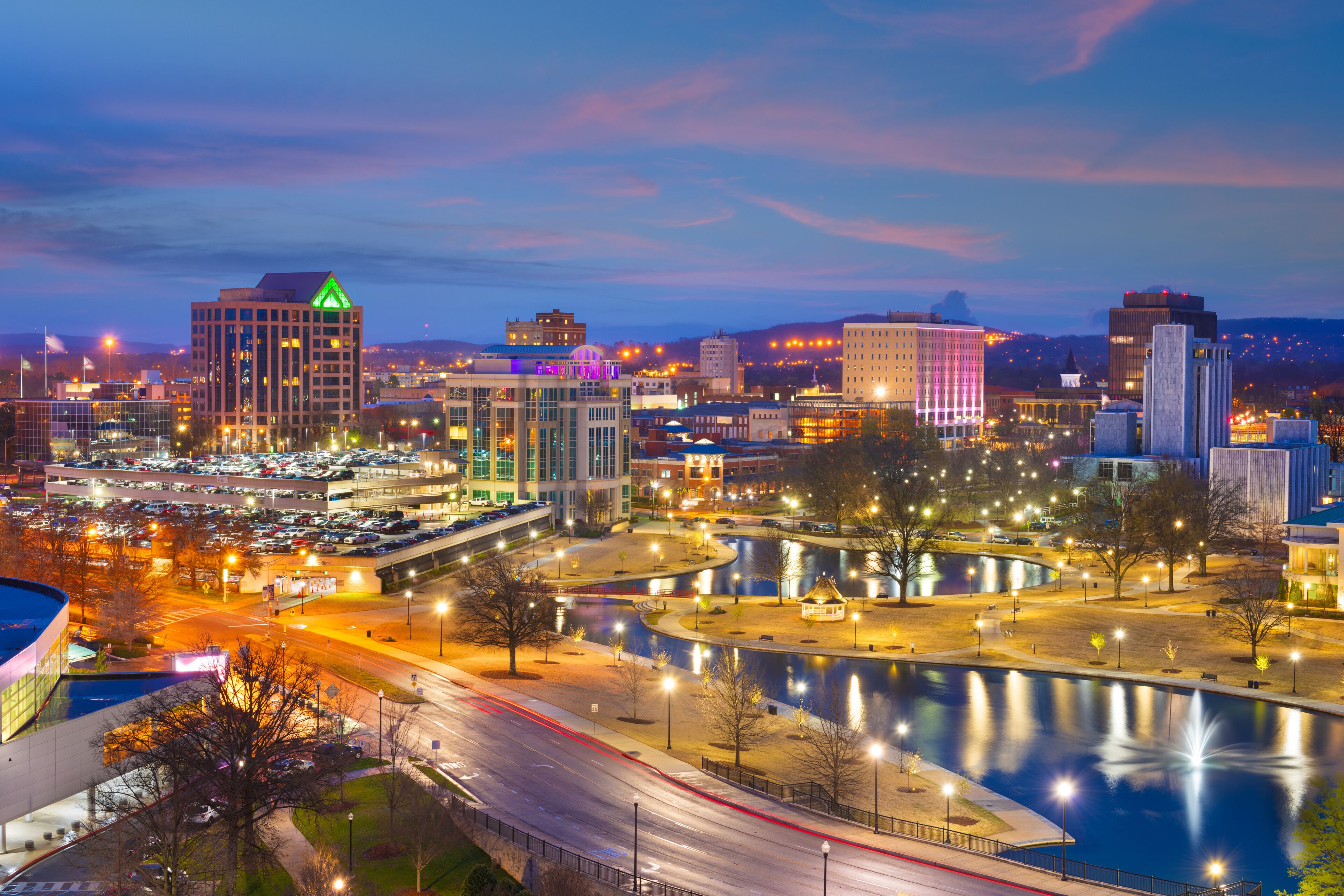 A cityscape at dusk with illuminated buildings, a river, and a bridge.