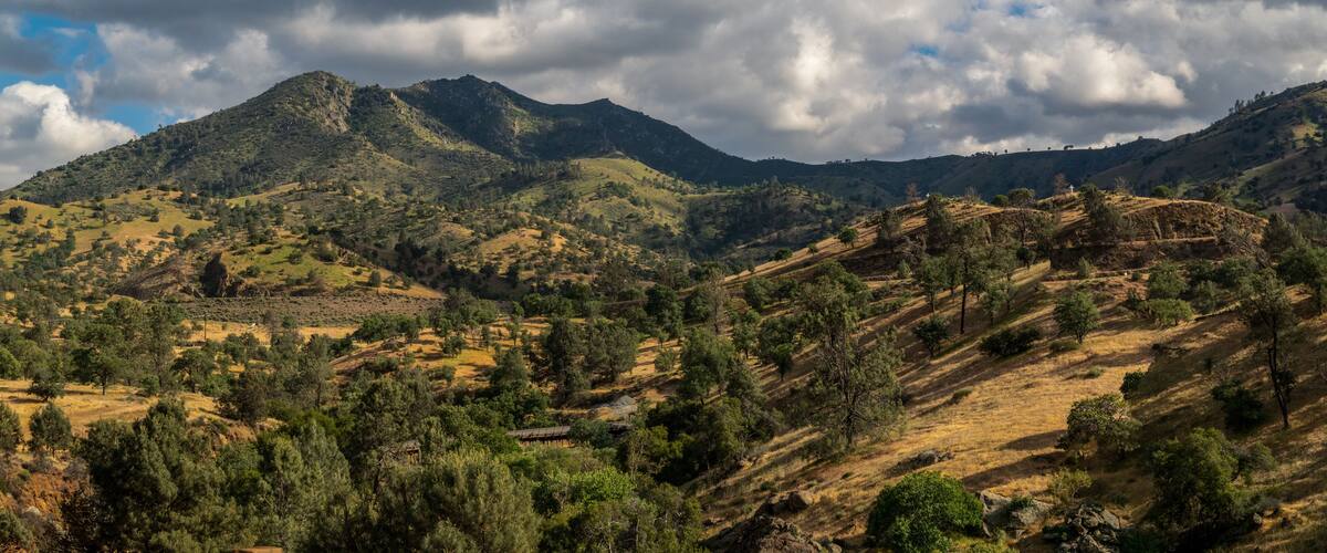 A mountainous landscape with scattered trees and a cloudy sky.