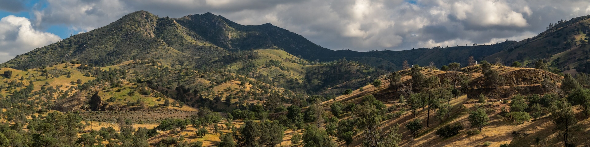 A mountainous landscape with scattered trees and a cloudy sky.