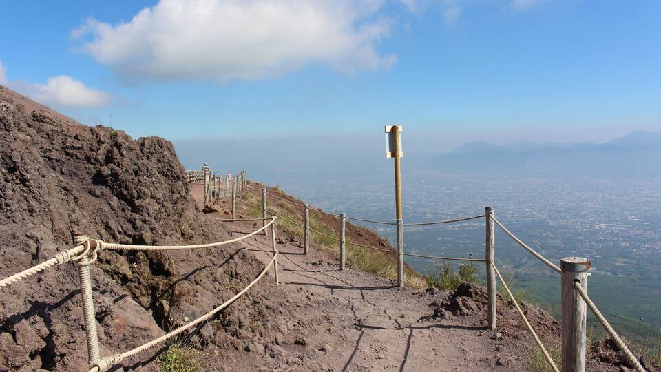 A rocky mountain path with a rope barrier, wooden posts, and a distant cityscape under a clear sky.